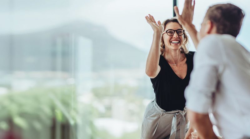 Image of two professionals high-fiving in an office in front of a large glass window to support roles of a good business manager article