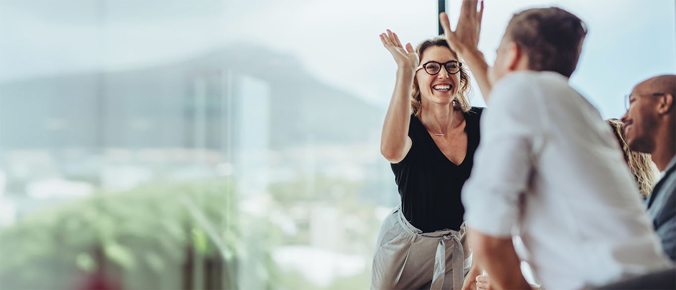 Image of two professionals high-fiving in an office in front of a large glass window to support roles of a good business manager article