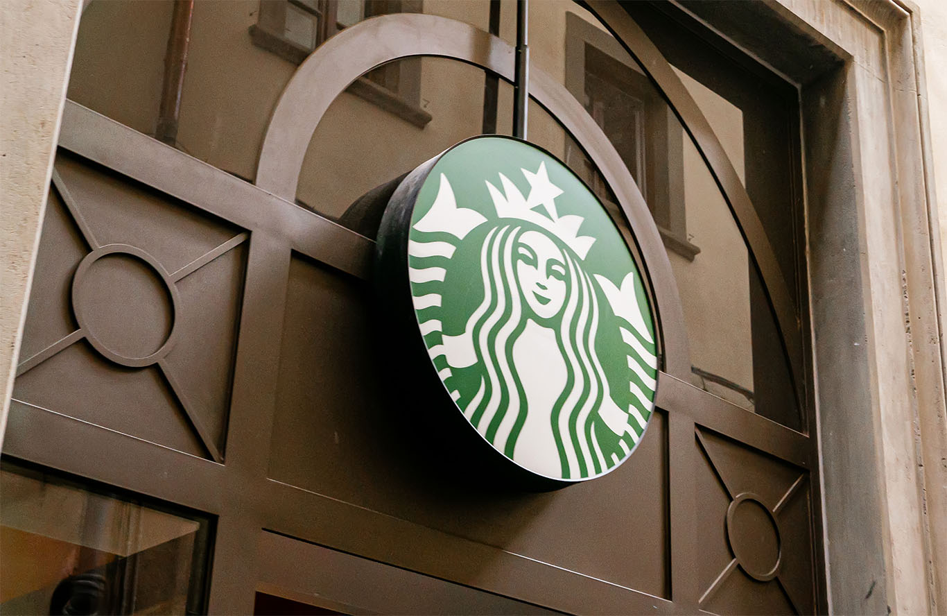 A Starbucks barista prepares a protein latte beside a digital order screen showing voice command features.