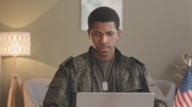 Military veteran wearing camouflage and dog tags using a laptop, with an American flag visible in the background.